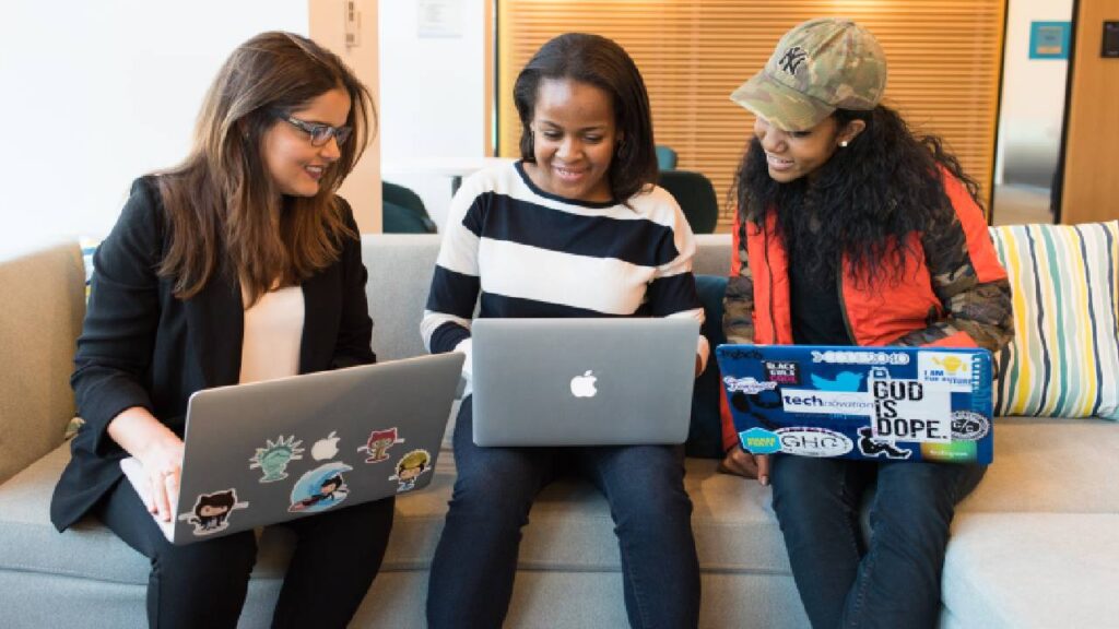 três mulheres sentadas à frente de computadores conversando sobre trabalho