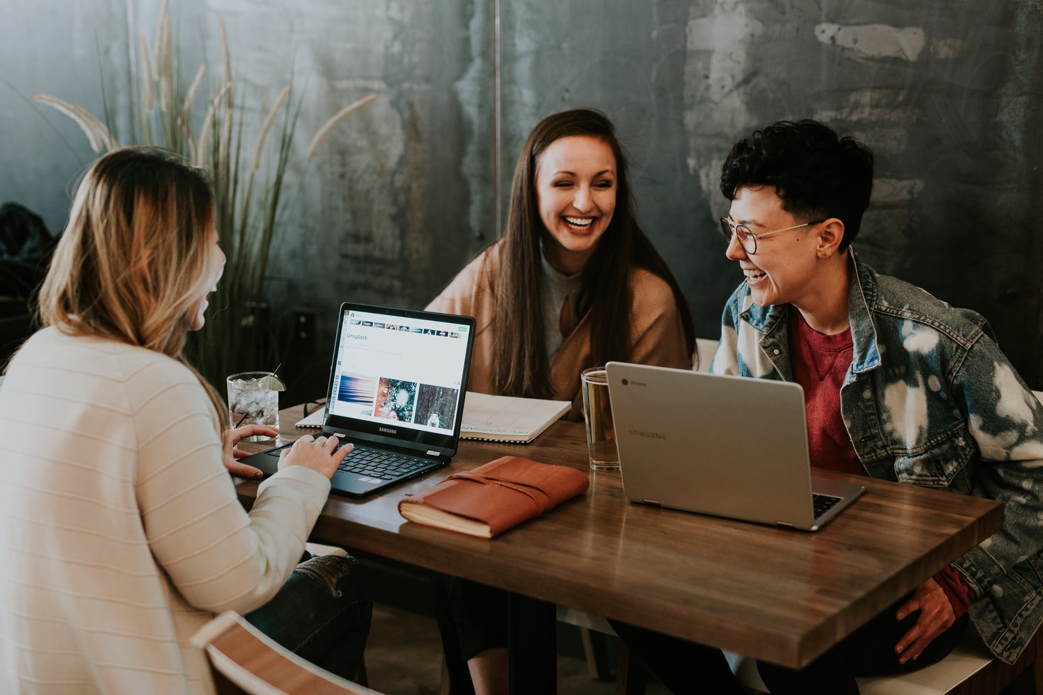 Vemos três mulheres sentadas à mesa; sorridentes, duas têm computadores a sua frente enquanto a terceira tem sobre a mesa um caderno (imagem ilustrativa).