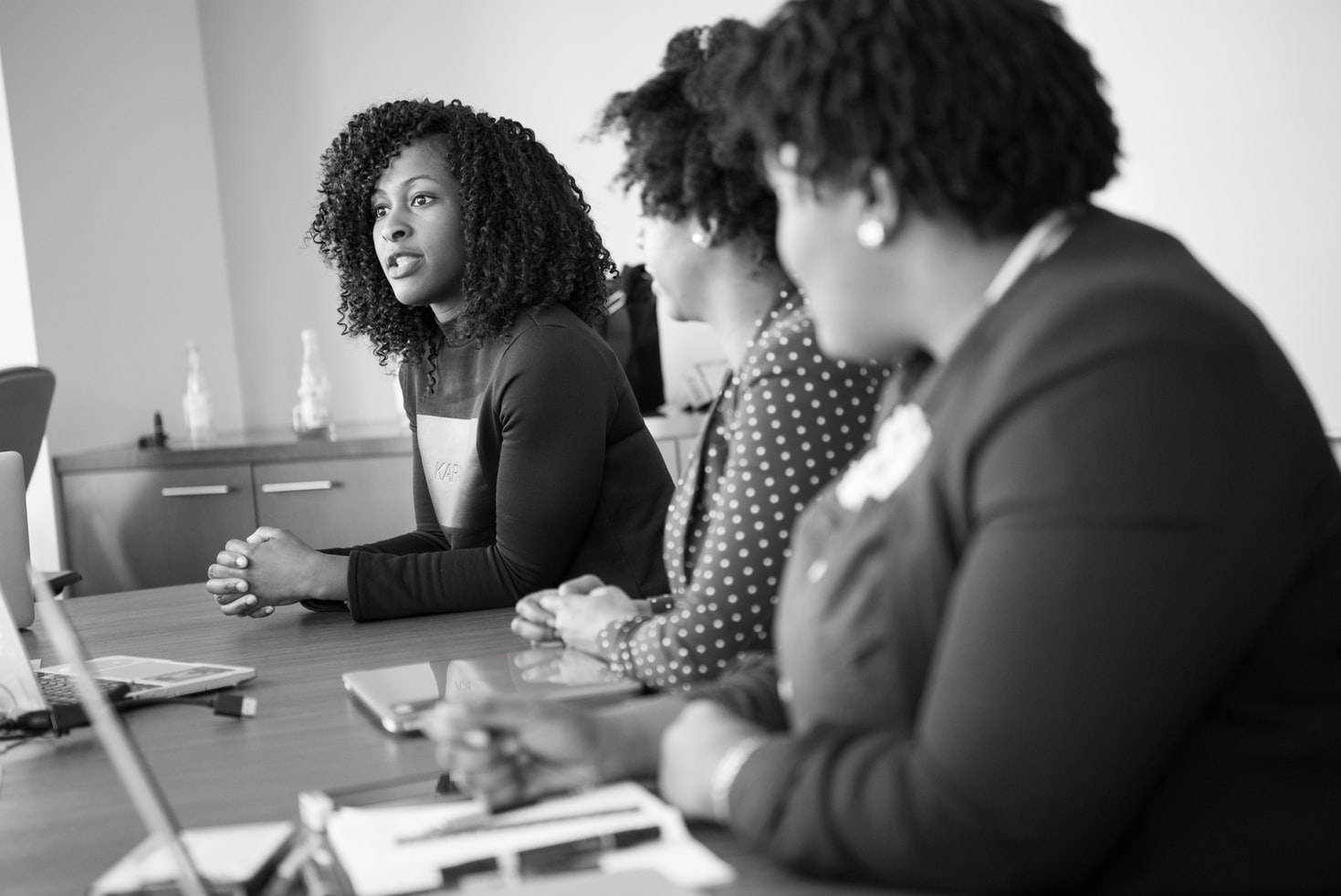 Vemos três mulheres negras sentadas à mesa; na ponta, umas delas tem a palavra. É possível ver, sobre a mesa, cadernos, canetas, computadores e outros acessórios (imagem ilustrativa). Texto: processo de inovação.