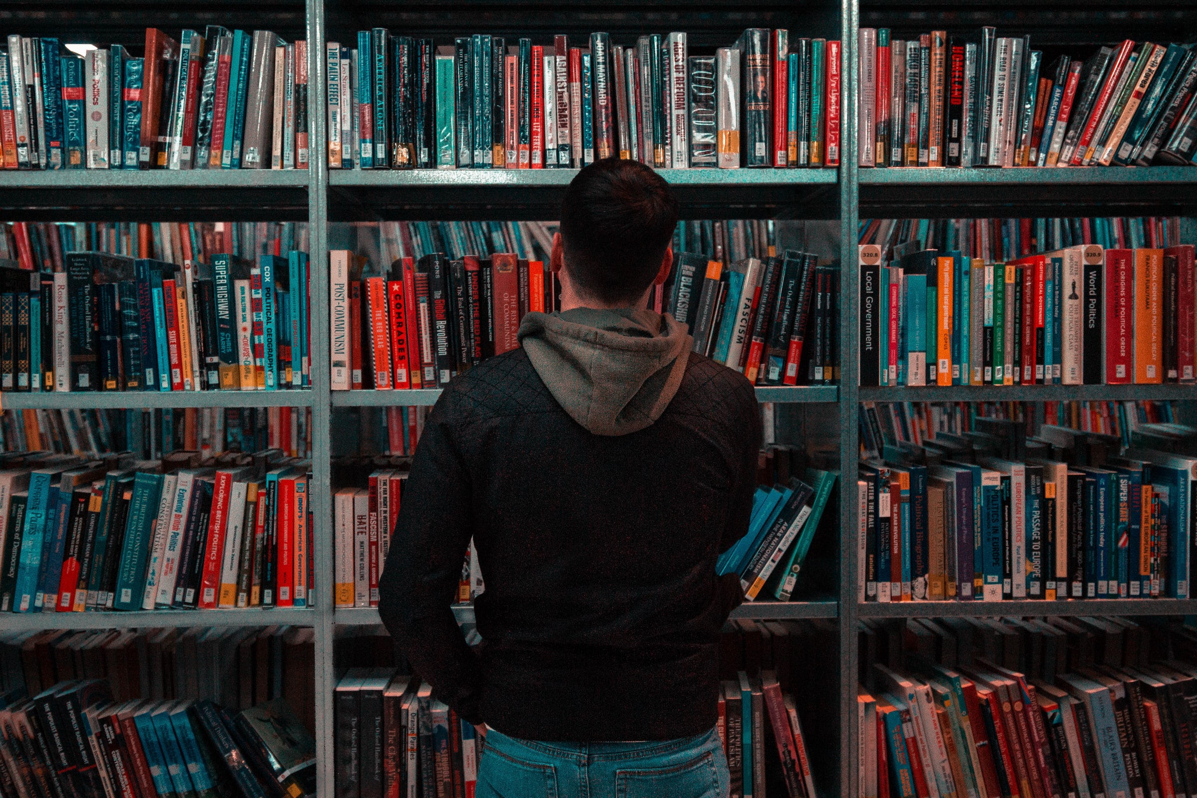 Vemos um homem parado em frente a uma grande estante de livros, no que parece ser uma biblioteca (imagem ilustrativa). Texto: gestão do conhecimento.