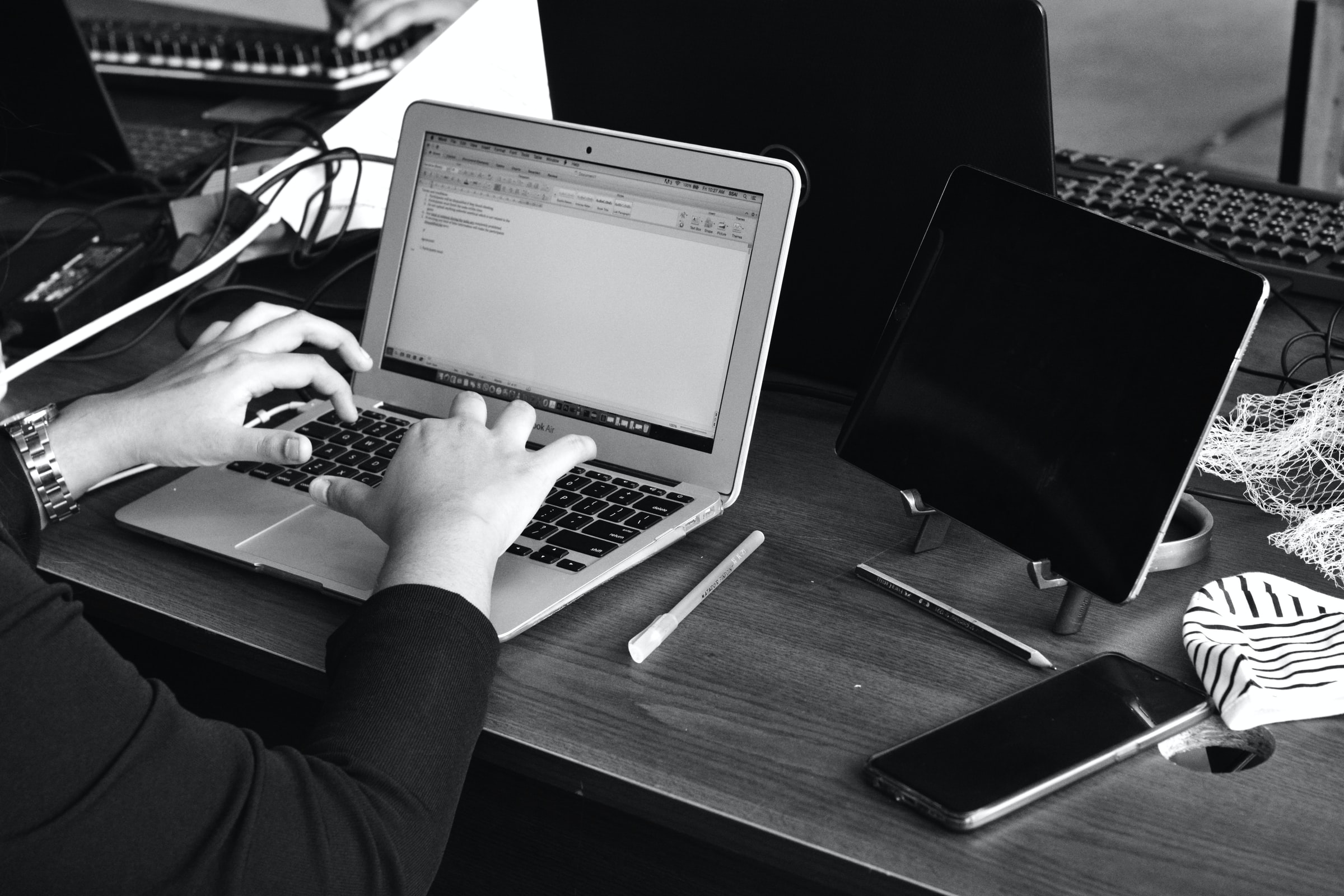 Vemos que uma pessoa tem sobre a mesa um computador, tablet, celular e canetas. Concentrada, ela trabalha em alguma tarefa (imagem ilustrativa).