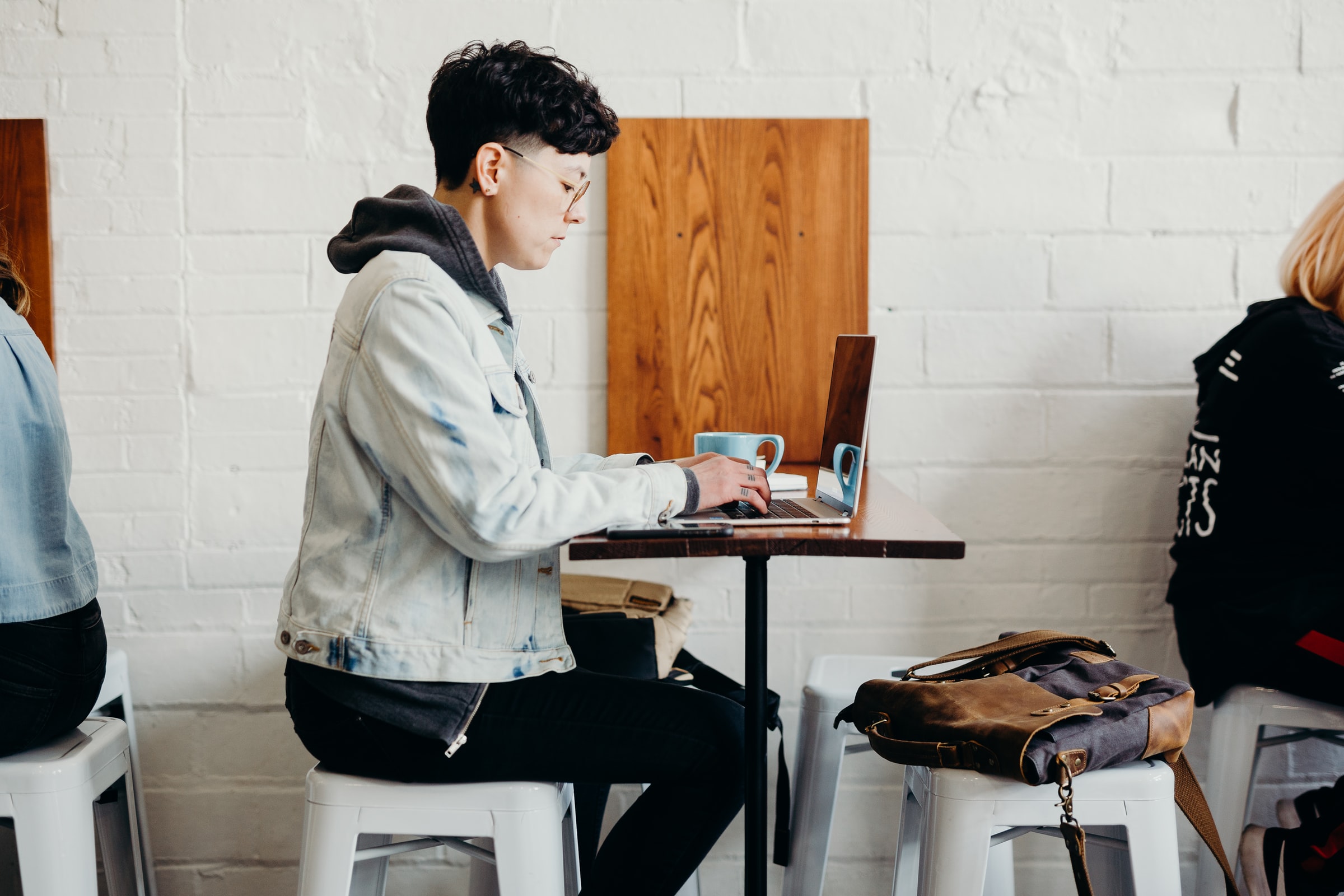 Vemos uma mulher trabalhando em um café. Sobre a mesa vemos uma caneca e seu computador (imagem ilustrativa).