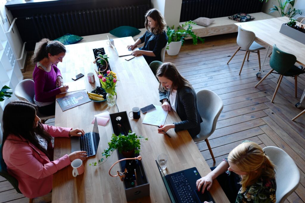Vemos uma mesa longa com 5 mulheres trabalhando sentadas. 3 com laptops e duas com folhas de papel à mesa.